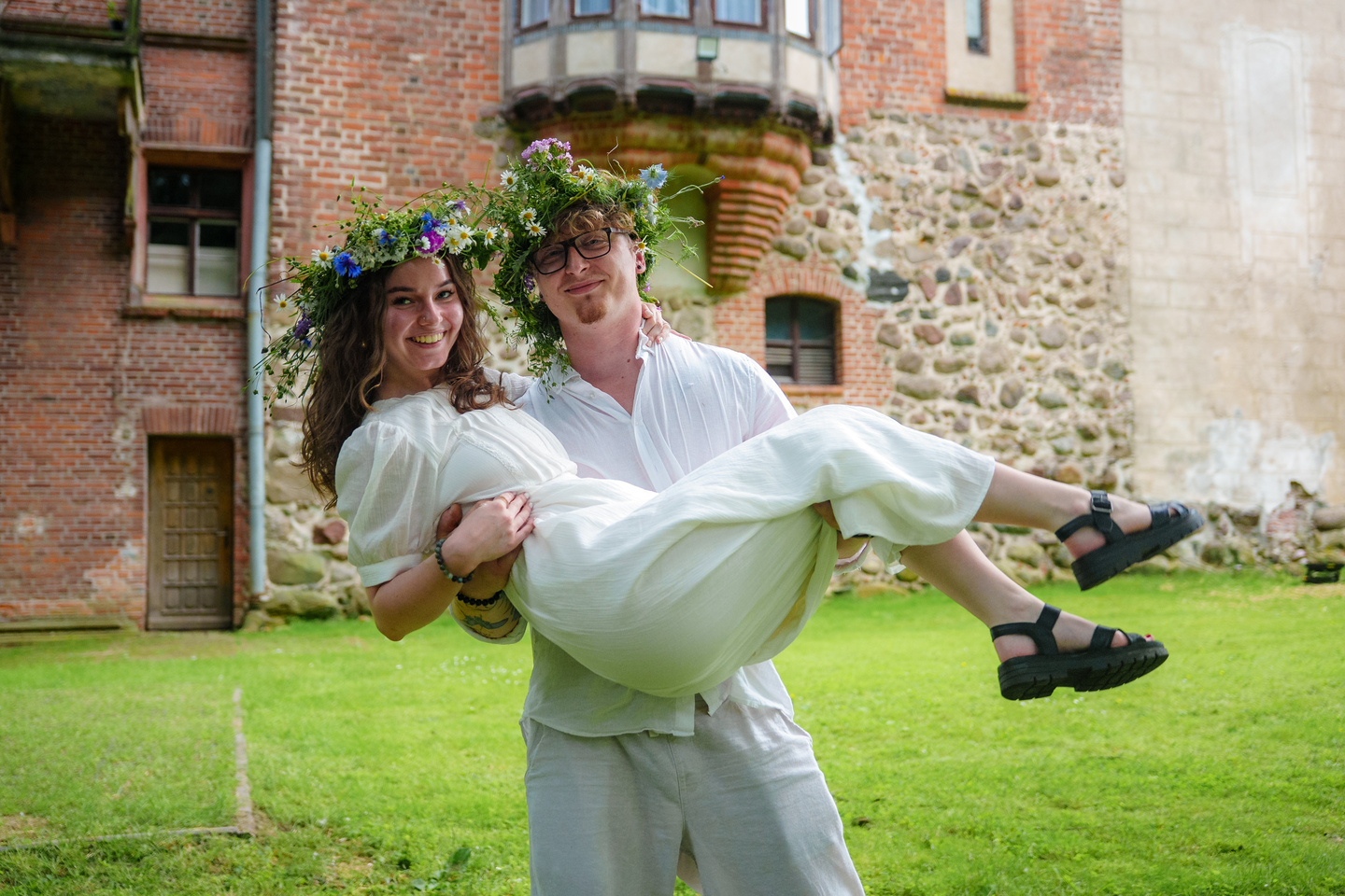 Couple in white outfits with flower crowns; man lifts smiling bride outdoors in front of a rustic brick building and grass.