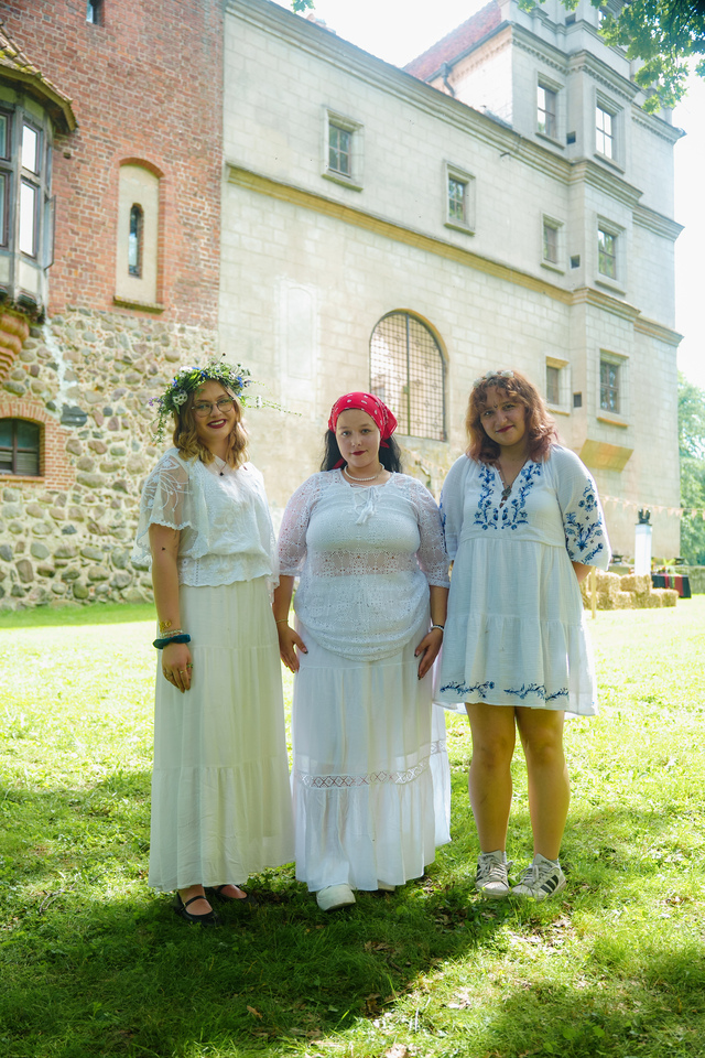 Three women in white dresses pose on a sunlit lawn, with a flower crown, red headscarf, and blue-embroidered outfit against a historic brick-and-stone building behind them.