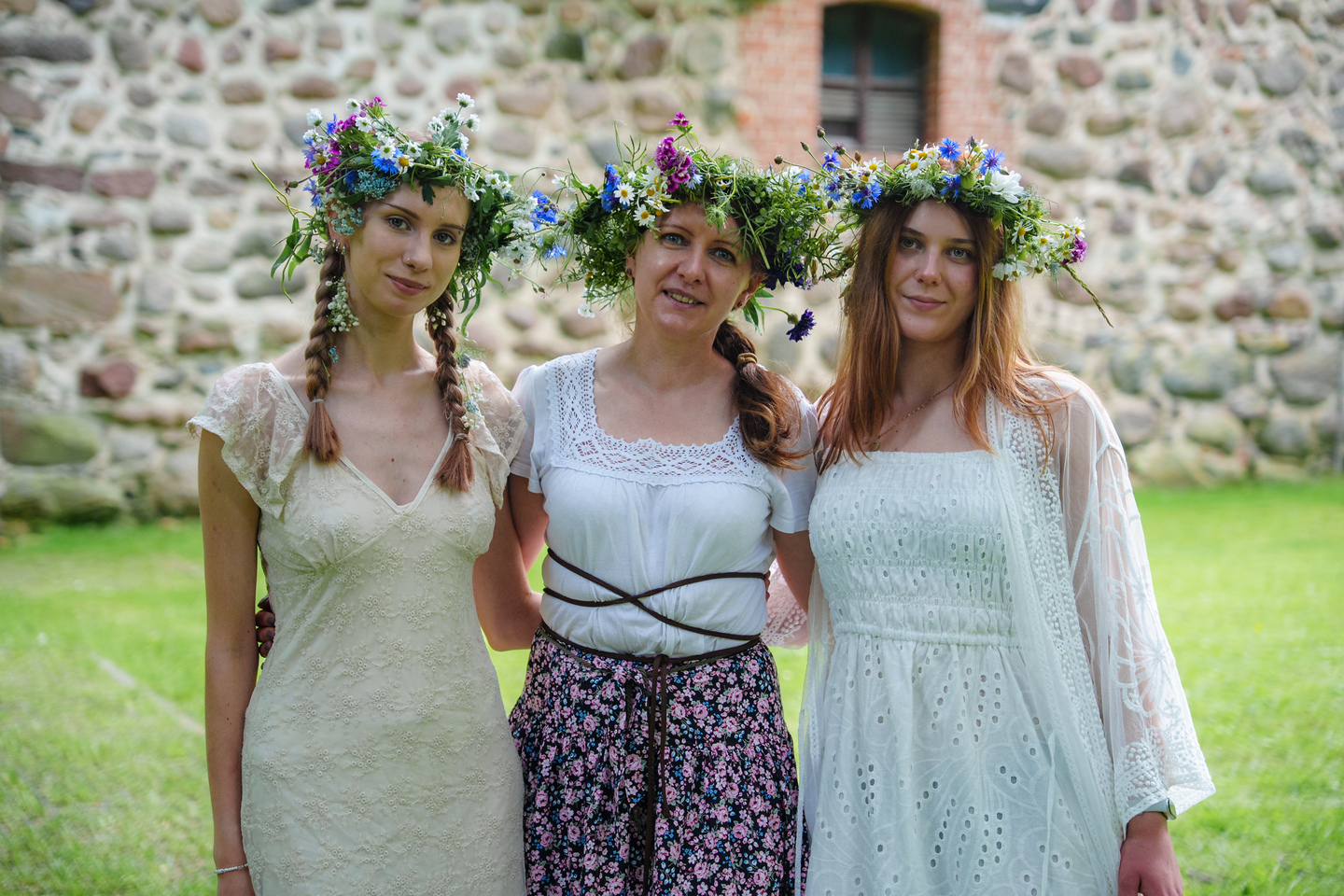 Three women wearing floral wreaths stand arm in arm on a lawn in front of a stone wall.