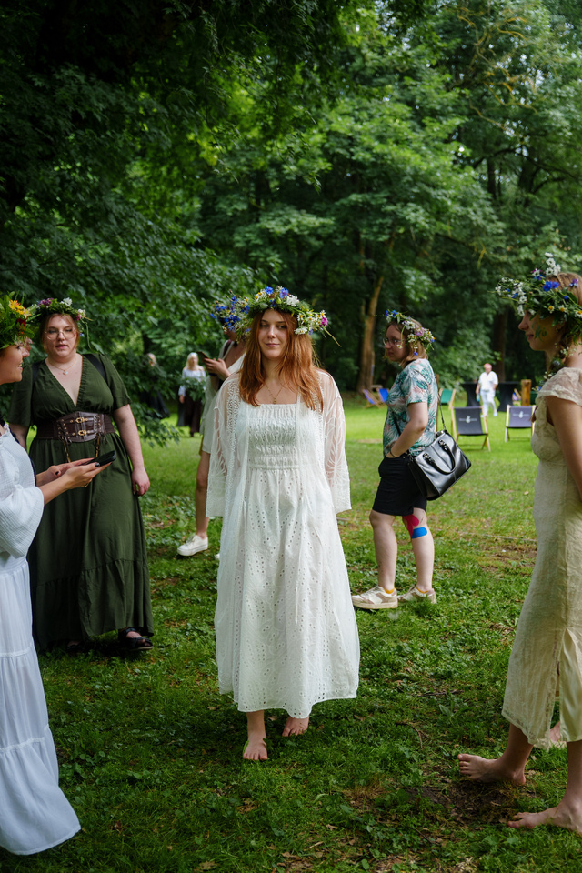 Young woman in a white dress with a flower crown stands barefoot on grass at an outdoor gathering in a park.