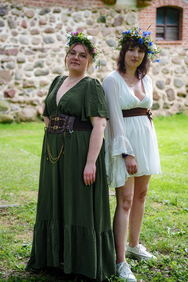 Two women in medieval-style dresses pose outdoors in front of a stone wall, wearing flower crowns and braided belts.