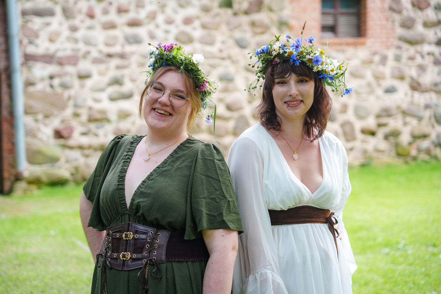 Two women in flower crowns pose outdoors in front of a stone wall, smiling; one wears a green dress with a corset belt, the other a white dress with a brown sash.