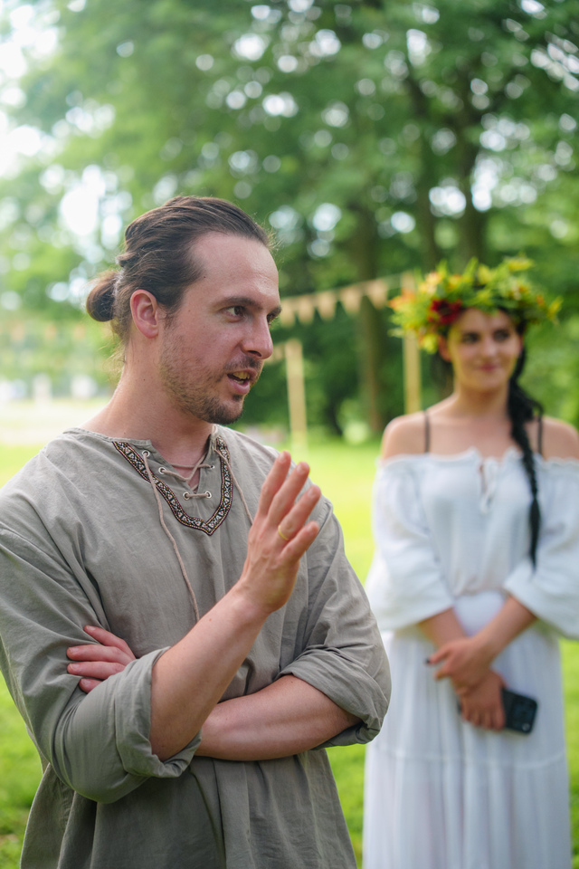 Man in a loose gray tunic gestures while speaking outdoors, with a woman in a white dress and flower crown standing in the background.