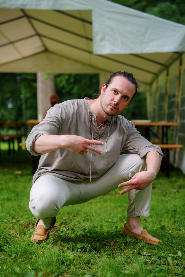 Man crouching on grass under a white event tent, making peace signs with both hands, wearing a beige tunic and light pants.