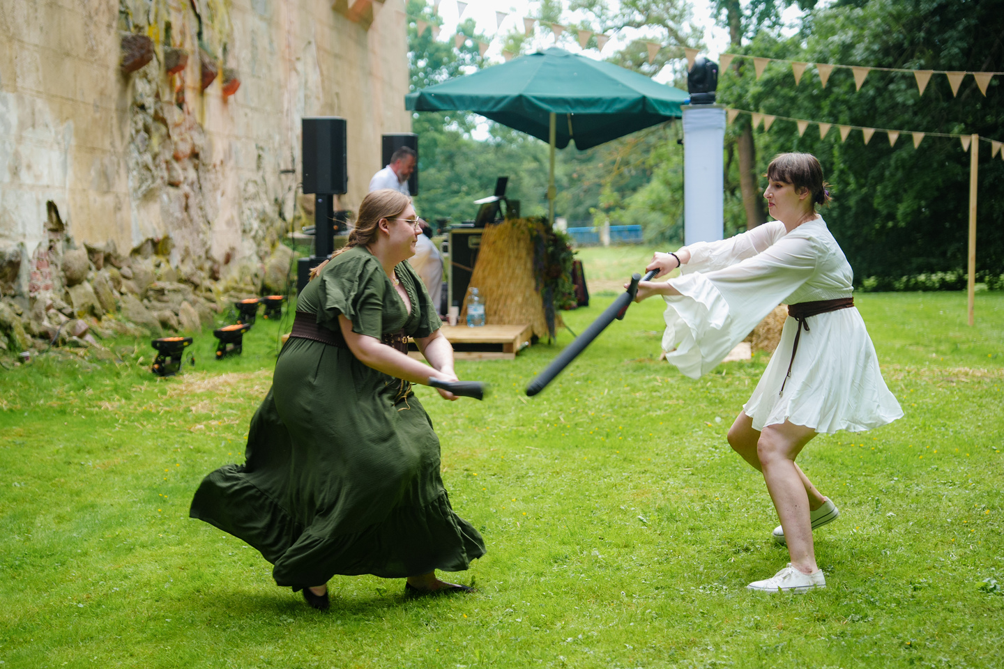 Two women in period costumes duel with foam swords on a grassy lawn, near a stone wall and festive bunting.