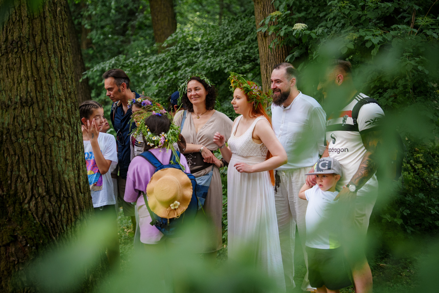 Group of people in a forest gathering, several wearing flower crowns and light, airy clothing, smiling and chatting.
