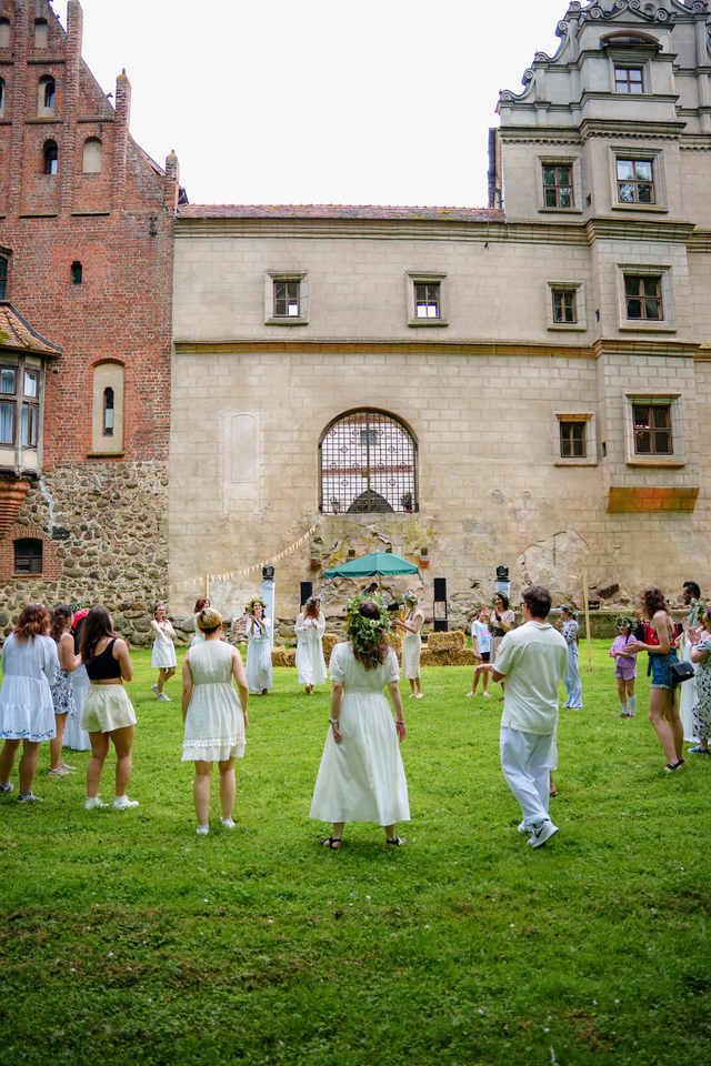 Group of guests in white dresses standing in a circle on a grassy lawn in front of a historic courtyard building.