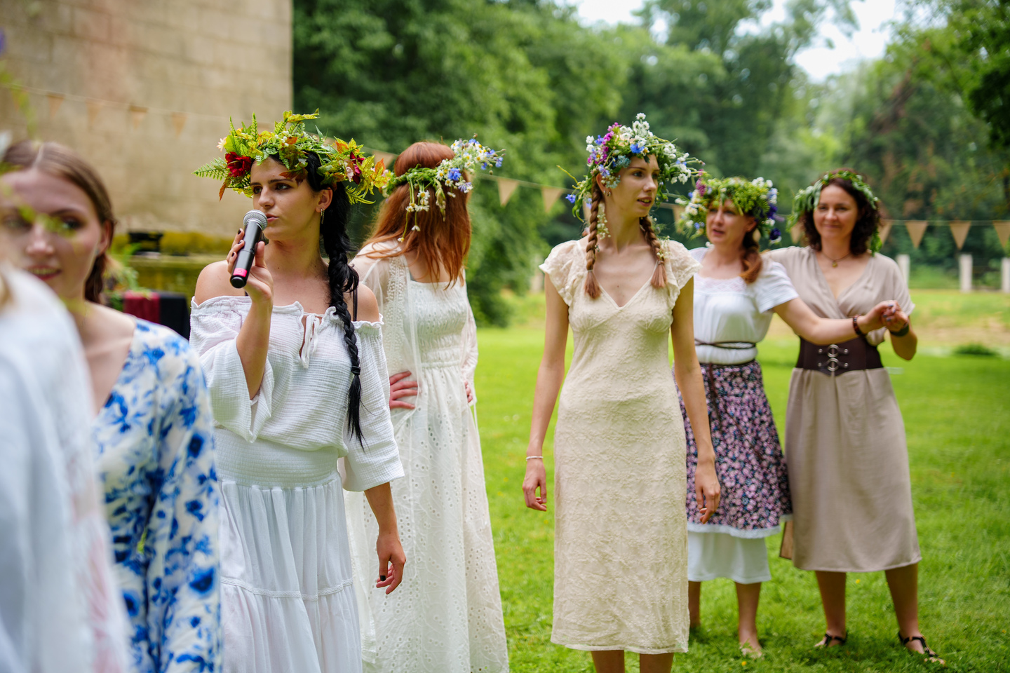 Women wearing flower crowns stand outdoors; one speaks into a microphone during a celebration.