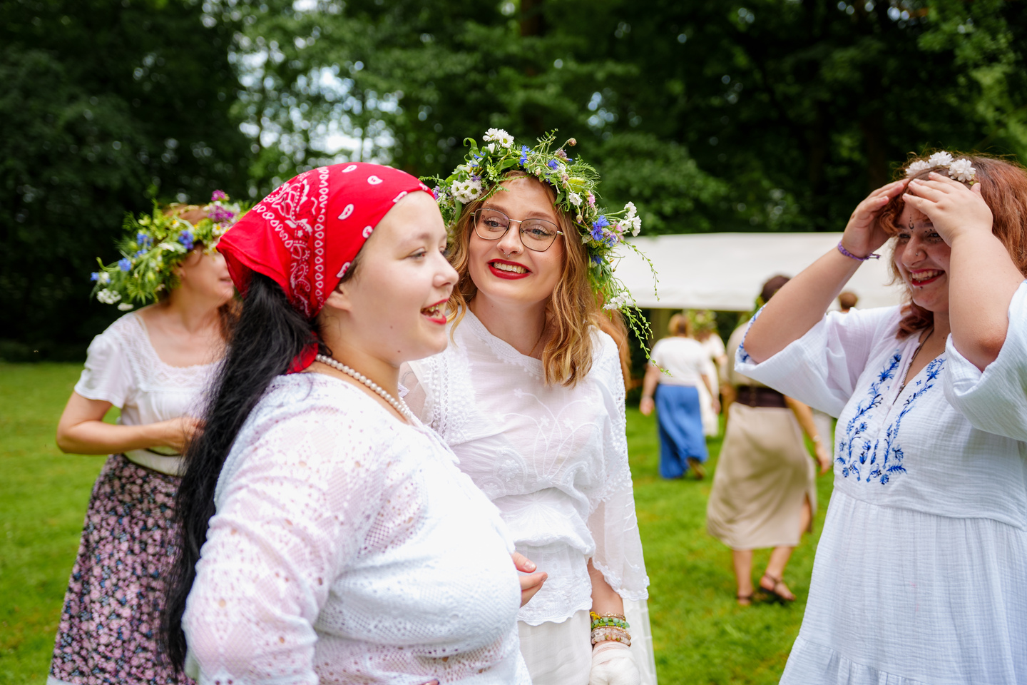 Women in white lace dresses wearing floral crowns laugh and chat at a sunny outdoor festival in a park.