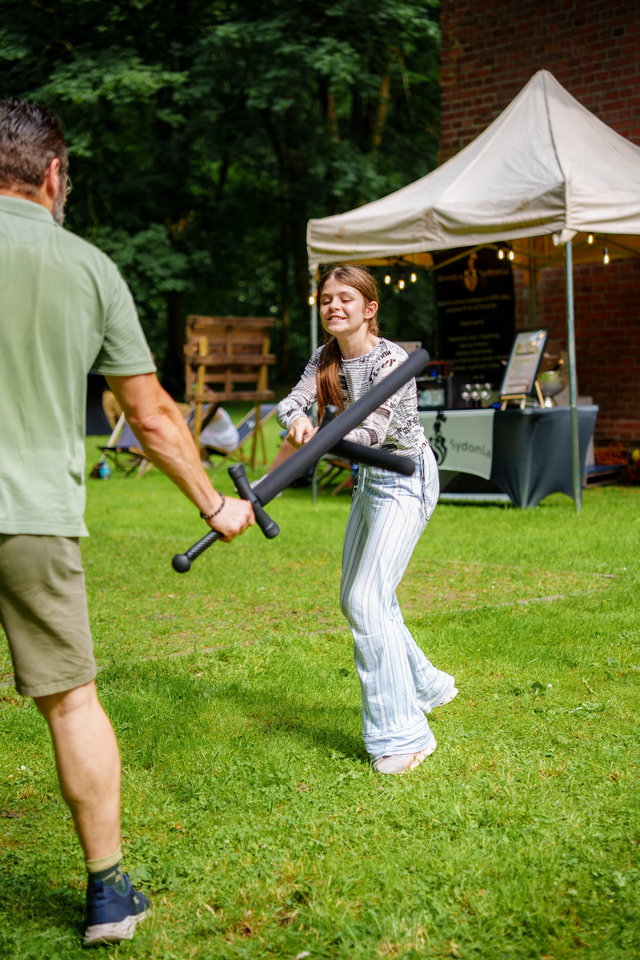 Young woman playfully swings a foam sword at a man during an outdoor festival, with a white tent and brick wall in the background.