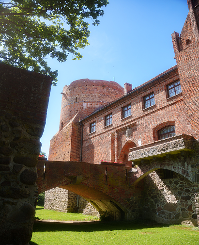 Brick medieval fortress with a round tower, stone arch bridge, and a green lawn under a blue sky.