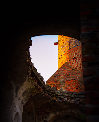 Sunlit brick tower visible through a crumbling stone arch in a ruined structure, rubble in the foreground.