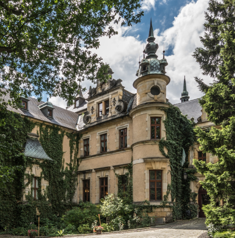 Historic manor with ivy-covered walls, a rounded turret, and decorative spires under a blue sky.