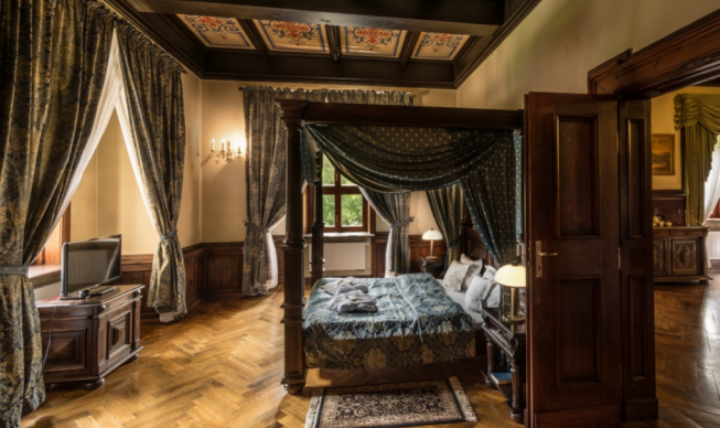 Luxurious four-poster bed in a classical hotel room with dark wood furniture, patterned drapes, and a parquet floor.