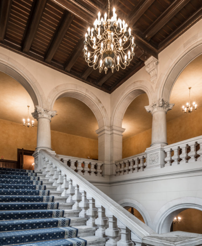 Grand marble staircase with a blue carpet, white balustrade, and arches in a historic mansion interior with a chandelier above.