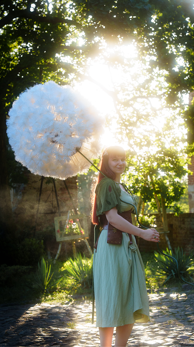 Young woman in a green dress stands in a sunlit garden, holding a large fluffy white dandelion-shaped parasol.