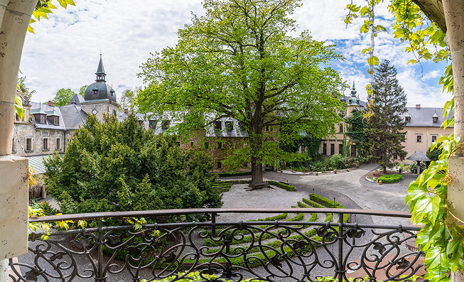Historic courtyard with stone buildings, lush trees, and a decorative wrought-iron railing seen from a balcony.
