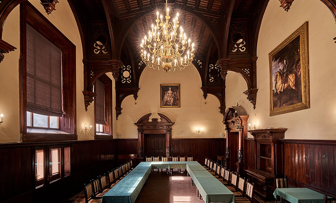 Elegant old-style conference room with dark wood paneling, a central chandelier, and a U-shaped table setup surrounded by chairs; portraits on the walls.