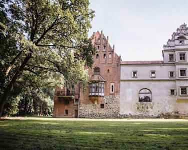 Ancient stone and brick castle wall with a small turret balcony, set on a grassy lawn with large leafy trees on the left.