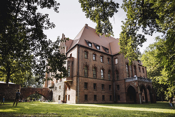 Brick castle-style brick building surrounded by trees on a sunny lawn in a park setting