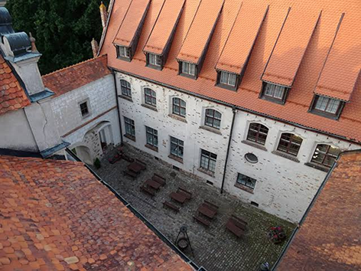 Aerial view of a white stone building with an orange tiled roof and a cobblestone courtyard lined with wooden tables and benches.