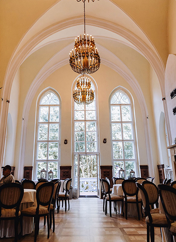 Elegant dining hall with arched ceilings, tall arched windows, and ornate chandeliers above rows of round tables and chairs.