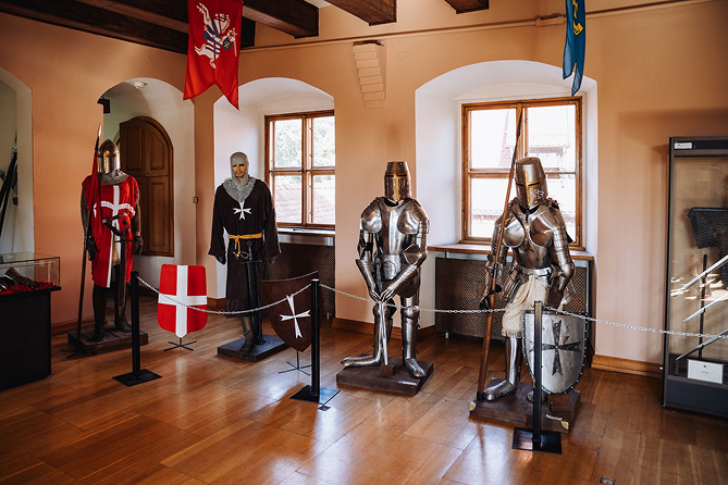 Museum display of medieval suits of armor with shields and banners, lined up along a wooden floor indoors