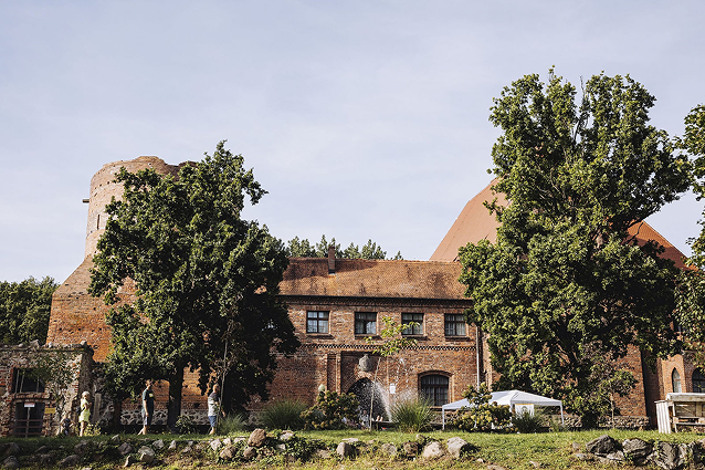 Brick historic building with a round tower, trees, and a water fountain in a grassy area outdoors.