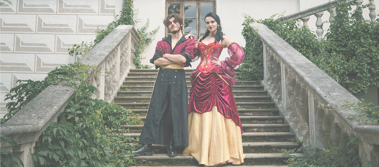 Two people in Renaissance-style costumes stand on a stone staircase covered in ivy, posing confidently.