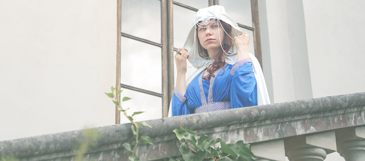 Woman in a blue medieval-style dress and white headscarf on a balcony, adjusting her veil.