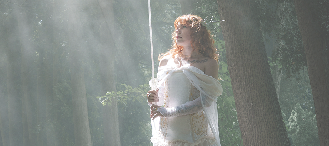 Woman in a white off-shoulder dress stands in a sunlit forest, holding a long sword upright.