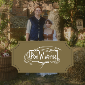 A young couple in medieval costumes stand together outdoors under a wooden arch, smiling at the camera with greenery and a rustic backdrop behind them.