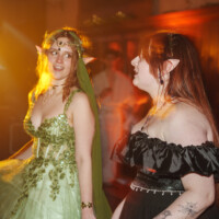 Two women in elf costumes at a glowing, warm-lit party, one in a gold-green beaded gown with a leafy headpiece and pointy ears, the other in a black off-shoulder dress with tattoos visible on her arm, both chatting or singing.