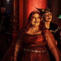 Two women in elaborate costumes pose together, one with a crown and elf ears, wearing a red dress under warm red lighting at a festive event.