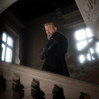A bearded man wearing a gold crown stands on a marble balcony in a dim, grand interior with arched architecture behind him.