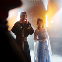 Two costumed performers on a foggy stage, backlit by bright spotlights and warm colors, smiling at the audience.