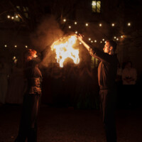 Two performers in dark clothing hold flaming props together, creating a bright fire between them at night under string lights.