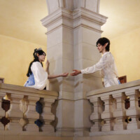 Two elf-like performers on a balcony, reaching toward each other, inside a stone-arched hall with beige walls and railing.