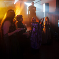 Performer in a dark costume on a stage with bright spotlights, while people in period dresses watch and clap nearby.