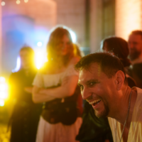 Man smiling in the foreground at an outdoor evening party, with colorful lights and other guests in the background.
