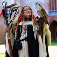 Woman in medieval fantasy costume with red dreadlocks, face paint, and fur cape, holding a decorated staff while a falcon perches on her gloved arm.