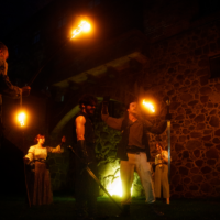 Group of performers in dark medieval costumes holding lit torches during a nighttime show in front of a stone wall.