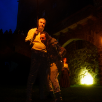 Two men in period costumes at night, one holding a blazing torch, with onlookers and a stone building in the background.