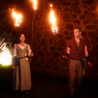 Two performers in medieval costumes hold flaming torches at night beside a stone wall, posing for a fire-performance scene.