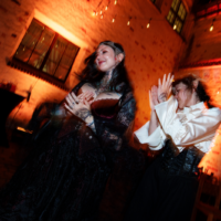 Two women in gothic attire dancing in a warm brick-walled venue; one with tattoos, the other in a white blouse and corset.