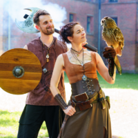 Couple in medieval costumes outdoors: man with shield and battle-ax stands beside a woman in a corseted dress, holding an owl on her gloved hand