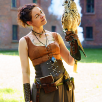 Woman in a brown medieval-style top and belt holds a perched owl on her gloved left hand outdoors.