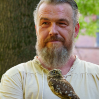 Bearded man in a light shirt outdoors with a small brown owl perched on his gloved hand.