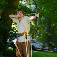 Archer in medieval-style clothing drawing a bow outdoors, arrows planted in a hay bale nearby under a leafy forest canopy