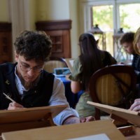 Two people sketch at wooden easels in a bright room, concentrating on their drawings while others work in the background.